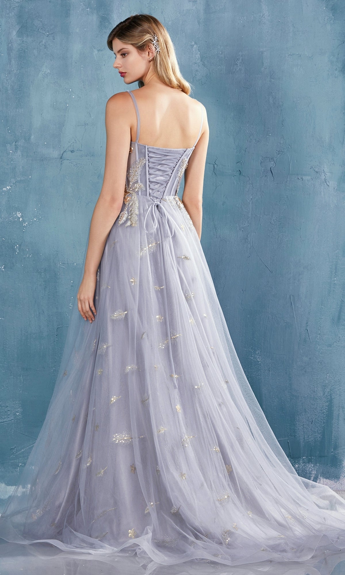 A woman stands against a blue textured wall, wearing the Shoulder-Bow Long Corset Prom Ball Gown A0824 with a corset bodice, thin straps, lace-up back, and gold embroidered details.